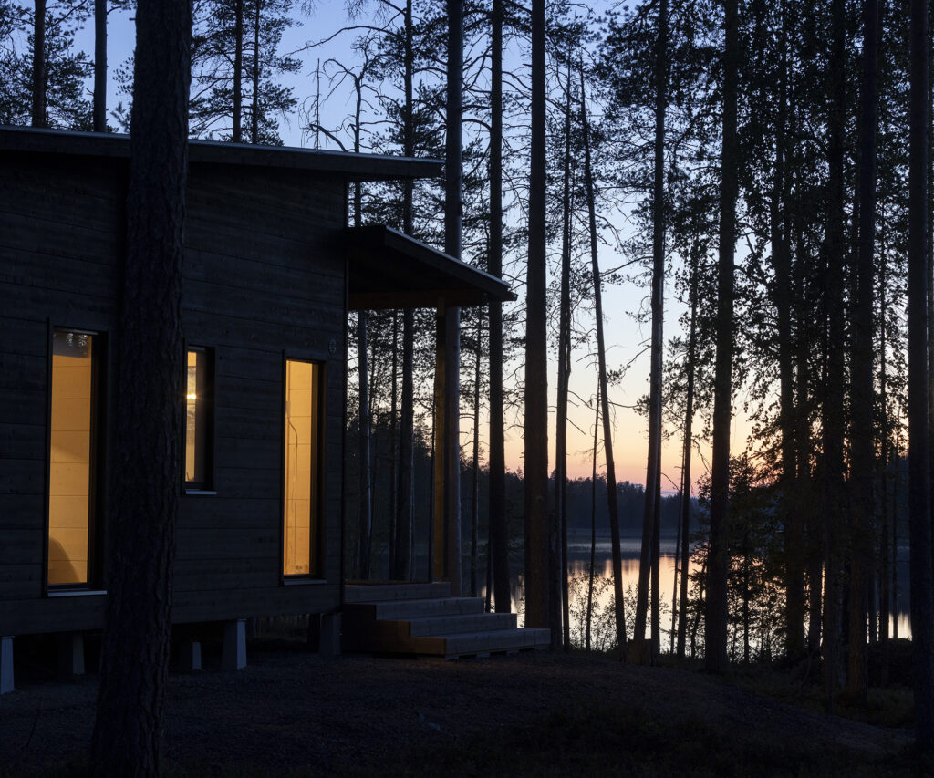 Mökin suurista ikkunoista näkyy valo. Tummuvassa illassa auringonlaskun kajo heijastuu puiden takana näkyvään järven pintaan.